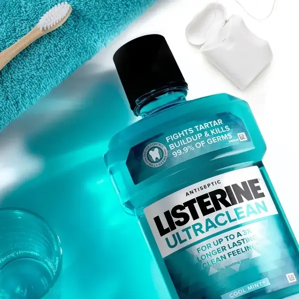 El producto Listerine Ultraclean Cool Mint apoyado sobre la superficie de un lavamanos. Un paquete de hilo dental, un cepillo de dientes, un paño y un vaso apoyados alrededor de la botella de Listerine.
