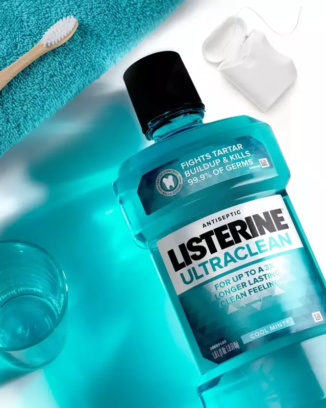 El producto Listerine Ultraclean Cool Mint apoyado sobre la superficie de un lavamanos. Un paquete de hilo dental, un cepillo de dientes, un paño y un vaso apoyados alrededor de una botella de Listerine.